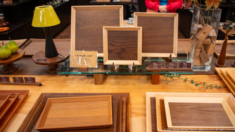 Wooden trays, shallow boxes, framed panels, and standing boards with varied grain patterns are arranged on a shop counter.