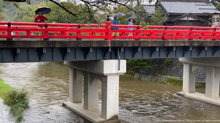 Bright red bridge over a brown river with pedestrians carrying umbrellas and trees along the bank.