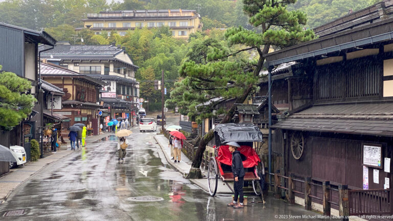 Rainy traditional street with dark wooden buildings, umbrellas, pruned pines, and a red rickshaw at right.