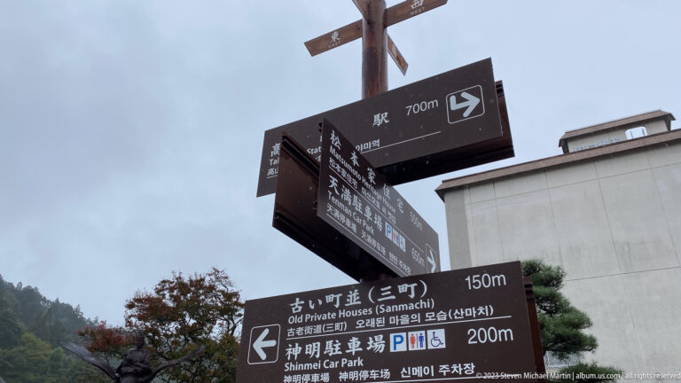 Brown multilingual directional signs point toward old private houses, parking, and other destinations under a gray sky.