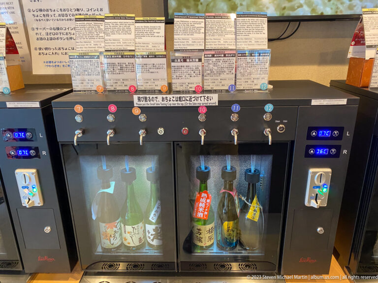 Self-service sake dispenser with six numbered taps, tasting notes, coin controls, and bottles chilled behind glass.