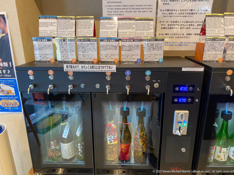 Wide view of adjoining sake dispensers with numbered taps, tasting cards, payment controls, and assorted bottles.