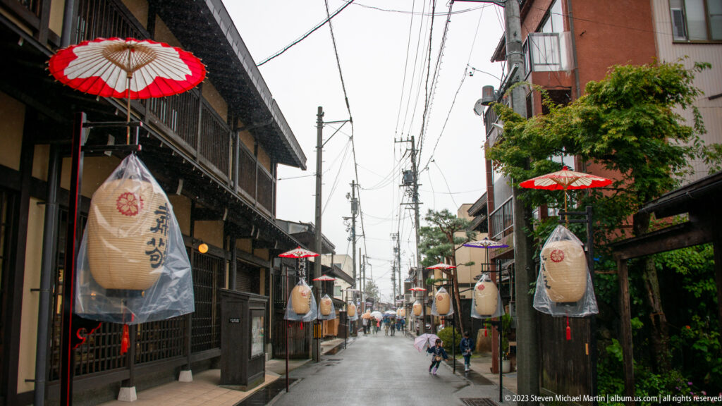 Rainy narrow street with dark wooden façades, hanging lanterns, red parasols, umbrellas, bicycles, and overhead utility lines.