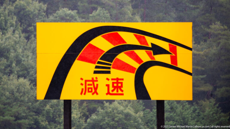 Yellow roadside sign with a black winding-road graphic and red Japanese characters meaning “slow down,” set against forested hills.