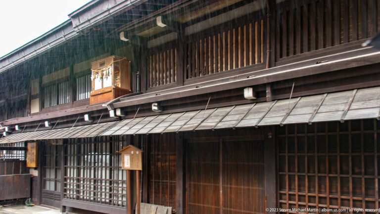 Dark timber townhouse façade with upper vertical slats, deep eaves, rain shelters, and ground-floor wooden lattice screens.