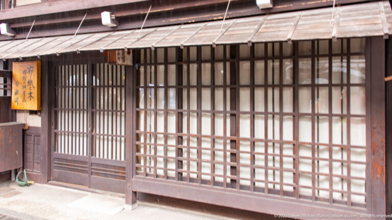 Traditional storefront with fine wooden lattice, translucent sliding panels, a weathered awning, and a projecting wooden sign.