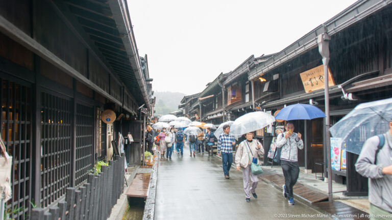 Umbrella-carrying visitors walk along a wet street lined with dark traditional wooden buildings and shop signs.