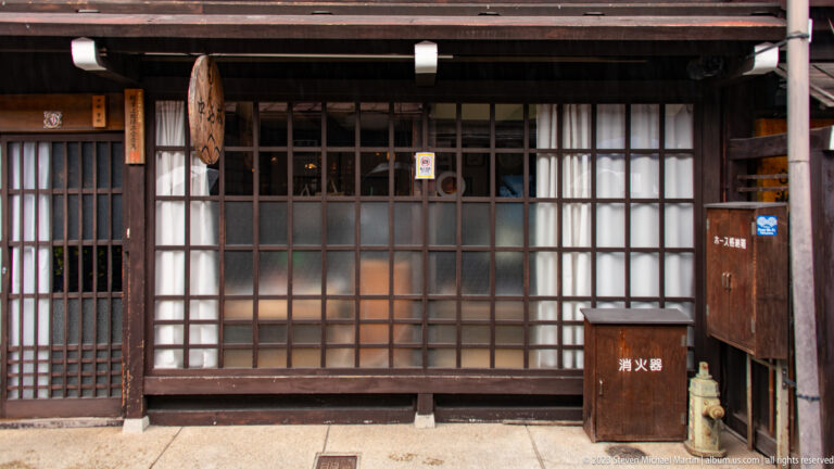 Dark timber storefront with gridded translucent glass panels, sliding doors, and a fire-extinguisher box at street level.