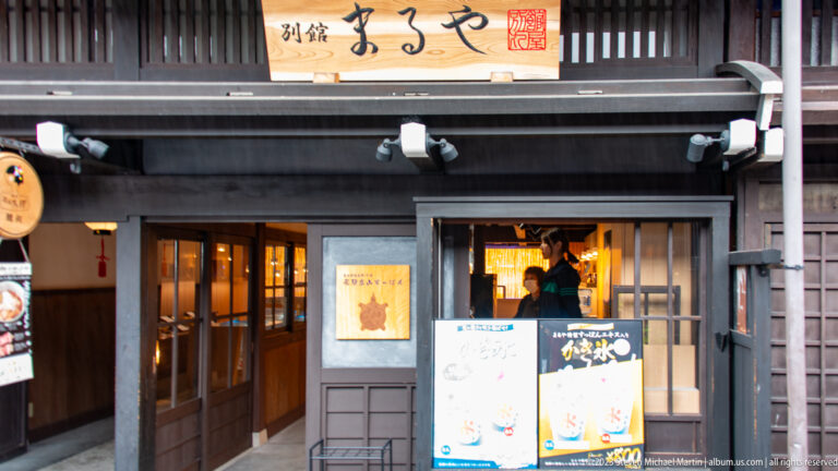 Dark wooden restaurant storefront with a large signboard, illuminated doorway, menu placards, and warm interior light.