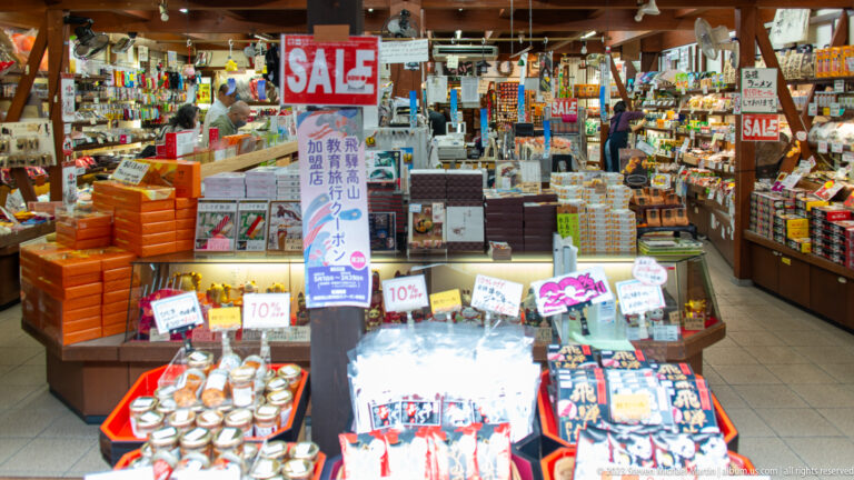 Crowded souvenir shop entrance filled with boxed foods, jars, gifts, postcards, price signs, and red sale banners.