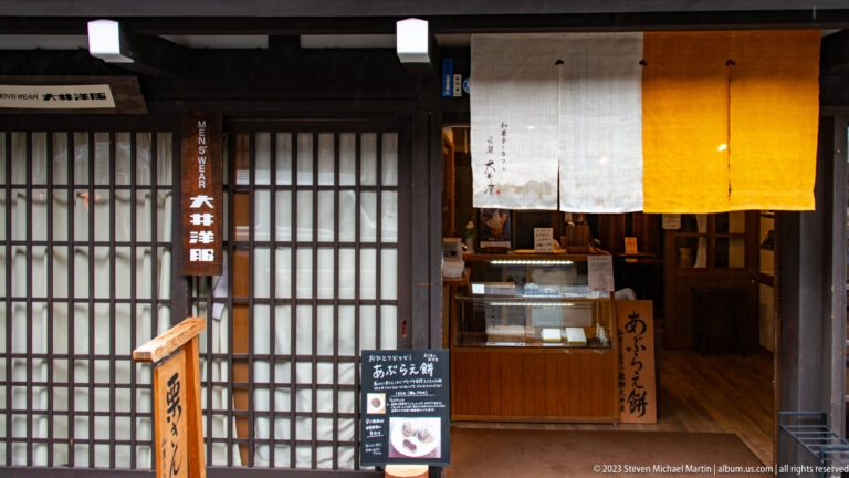 Wooden confectionery storefront with white-and-yellow noren curtains, a sweets sign, lattice doors, and a small display counter.
