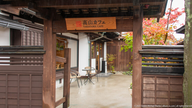 Timber passage labeled Takayama Cafe leads into a small courtyard with outdoor tables and autumn foliage.