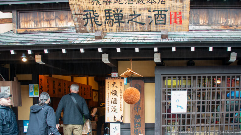 Visitors enter a traditional sake shop beneath a large sign and hanging cedar sugidama, with bottles and calligraphic boards inside.