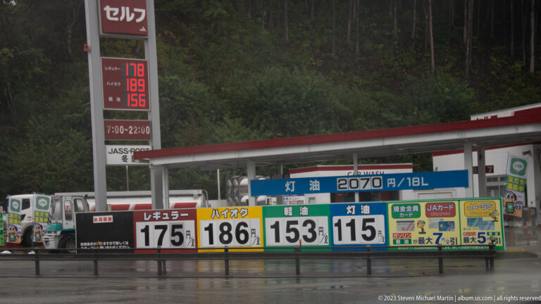 Japanese fuel station price board lists several fuel grades beneath a red canopy beside a wet mountain road.