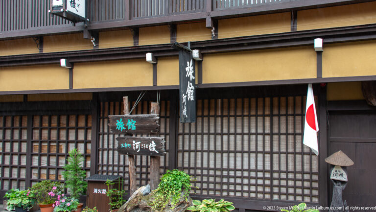Traditional wooden lodging façade with lattice screens, small garden, stone ornament, vertical signs, and a Japanese flag.