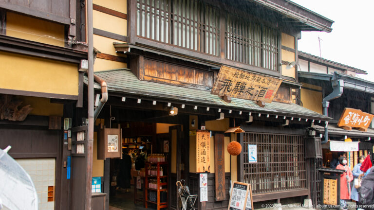 Traditional timber sake storefront with a weathered signboard, hanging cedar sugidama, dark latticework, and vertical calligraphy.