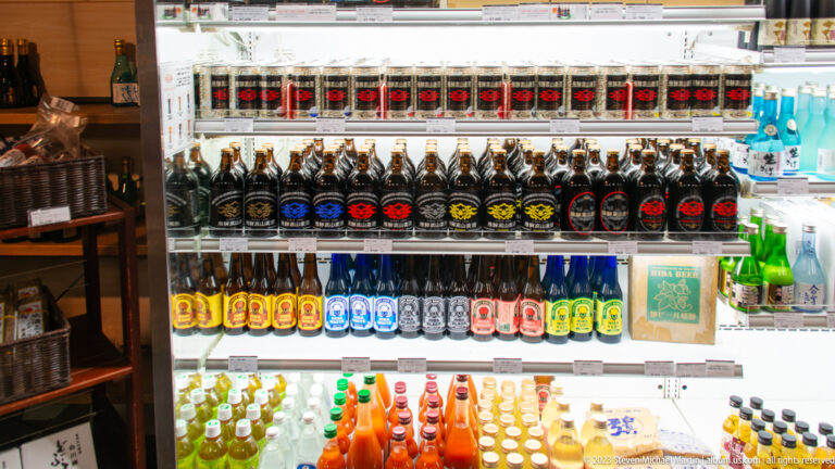 Bright refrigerated case stocked with rows of dark sake bottles, colorful drinks, and packaged goods on lower shelves.