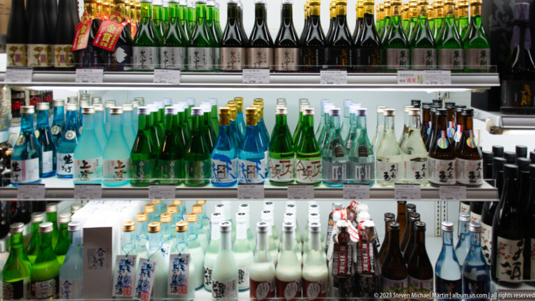 Refrigerated shelves hold clear, green, blue, and dark Japanese sake bottles with varied labels and caps.