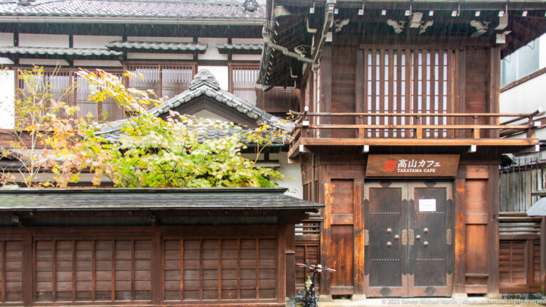 Tall traditional timber gatehouse with deep eaves, upper slats, heavy doors, and a Takayama Café sign at street level.