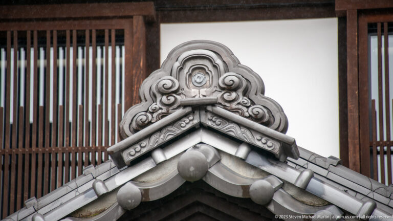 Gray ornamental roof-end tile with layered scrollwork, round bosses, and a central crest above intersecting roof slopes.