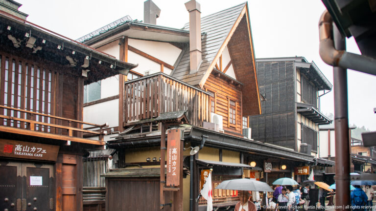 Steep-gabled wood-clad building rises behind traditional timber shopfronts and umbrellas on a Takayama street corner.