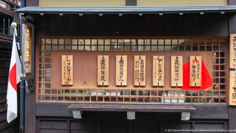 Row of handwritten wooden Japanese menu plaques mounted on a dark lattice storefront beside a Japanese flag.