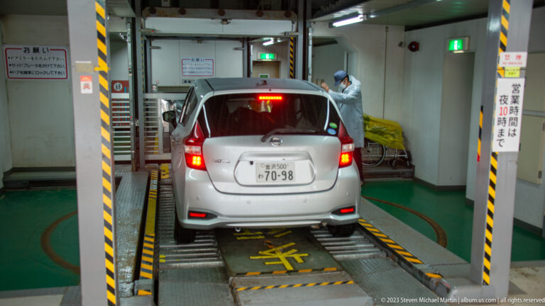 Compact car sits on a metal guided platform inside a mechanical parking facility while an attendant stands near the rear.