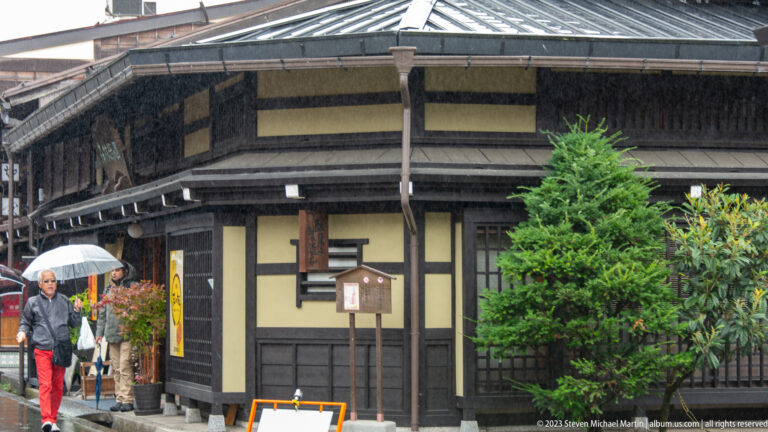 Dark timber townhouse corner with broad eaves, pale wall panels, potted greenery, and pedestrians carrying umbrellas.