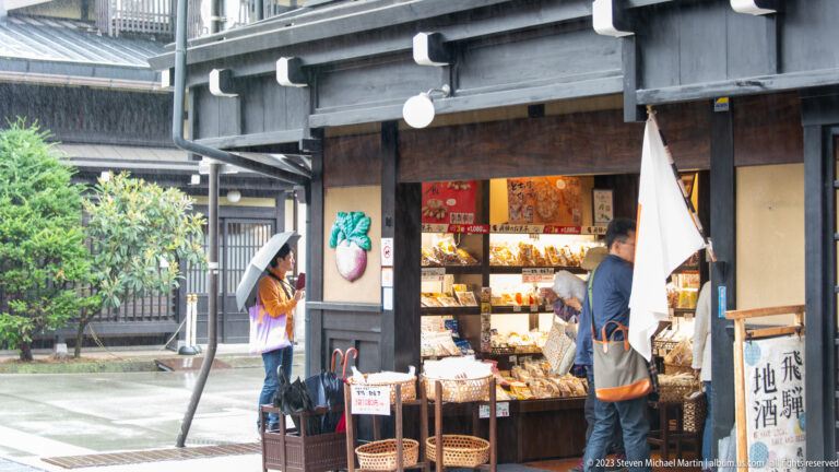 Customers browse a small open-front timber shop with baskets, packaged foods, a white hanging cloth, and local sake signage.