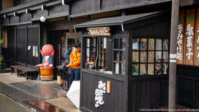 Small black-timber sweets shop with service window, seated customer, umbrella, and large red sarubobo figure outside.