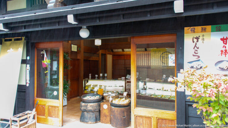 Open wooden confectionery shop with glass sweet display, barrel stands, flowering branches, and a vertical dango sign.