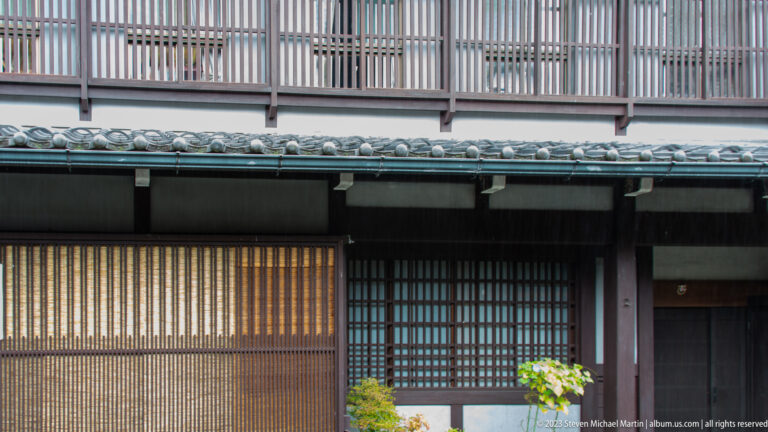 Two-story dark wooden townhouse with upper vertical slats, tiled eaves, and fine ground-floor lattice screens.