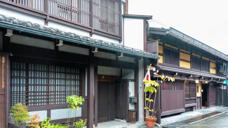 Adjoining traditional wooden townhouses with varied lattice patterns, potted plants, tiled eaves, and narrow street frontage.