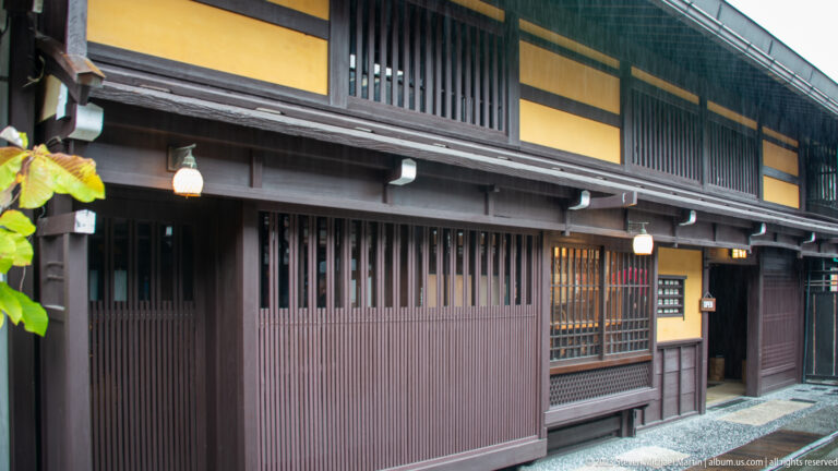 Long dark timber shopfront with vertical slats, deep eaves, yellow upper panels, lamps, and recessed entrances.