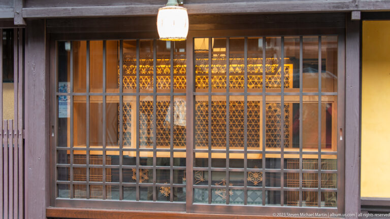 Wooden grid storefront window reveals warm illuminated geometric lattice panels inside beneath a hanging paper lantern.