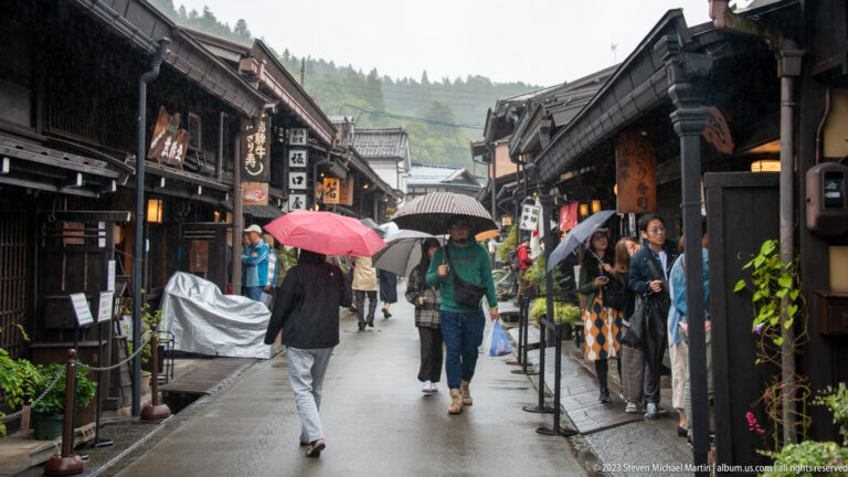 Visitors with umbrellas walk along a narrow wet street lined with dark timber shops, signs, and distant green hills.