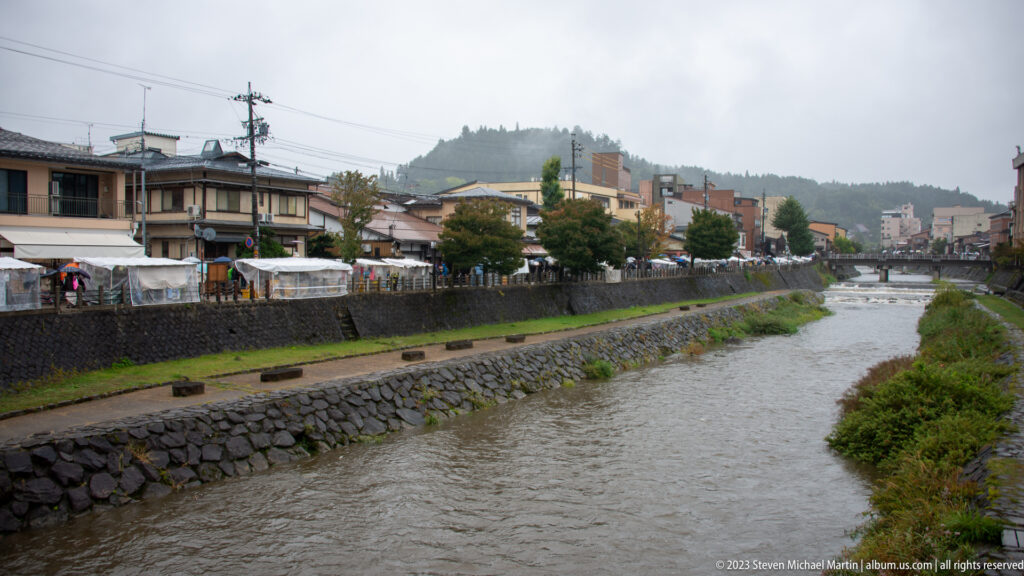 Shallow river flows between stone embankments through Takayama, with low buildings on one bank and wooded hills beyond.