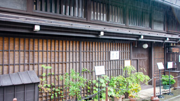 Traditional dark timber storefront with dense lattice screens, upper slats, potted plants, and small signs along the street.