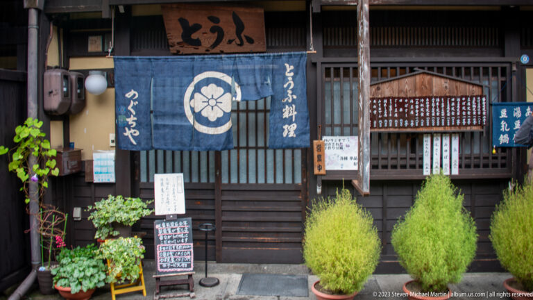 Traditional tofu restaurant with blue floral-crest noren, Japanese signs, dark timber lattice, menu boards, and clipped shrubs.