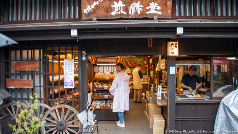 Customers browse a traditional open-front food shop with prepared items, hanging packages, side counter, and wooden cart wheel.