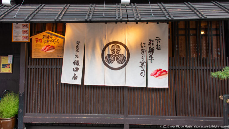 White noren with circular crest and Japanese calligraphy hangs across a dark wooden restaurant entrance beside a red food motif.