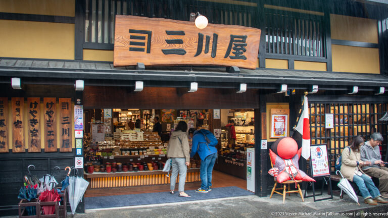 Miyagawa Morning Market storefront with large wooden sign, browsing customers, Japanese flag, bench, and red sarubobo figure.