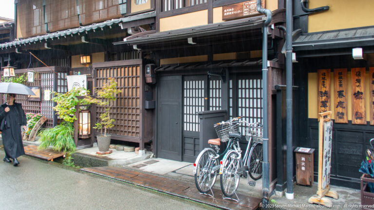 Dark timber shopfront with lattice screens, potted plants, two parked bicycles, wet pavement, and an umbrella-carrying pedestrian.