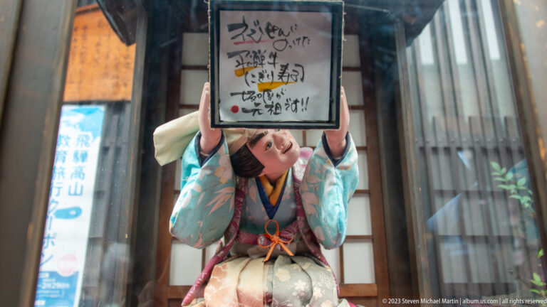 Woman in layered historical-style Japanese dress holds a handwritten sign overhead beside a reflective shop window.
