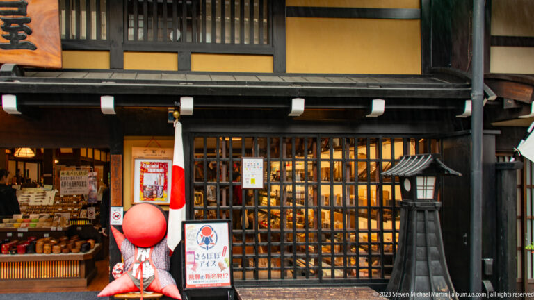 Large red sarubobo figure stands beside a Japanese flag at an open market shop filled with jars and packaged goods.
