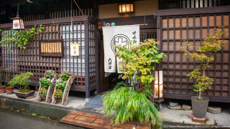 Traditional restaurant doorway with white noren, dark latticework, glowing lantern, potted maple, grasses, and flowers.