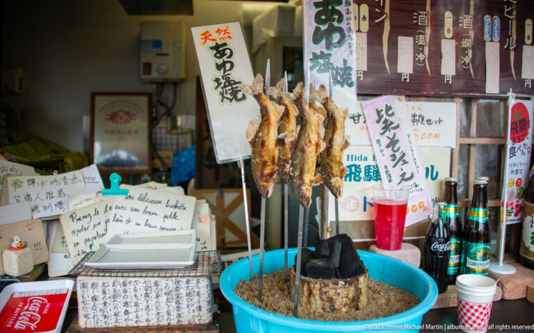 Skewered whole fish stand around a small charcoal hearth amid handwritten signs, bottles, and serving vessels in a shop.