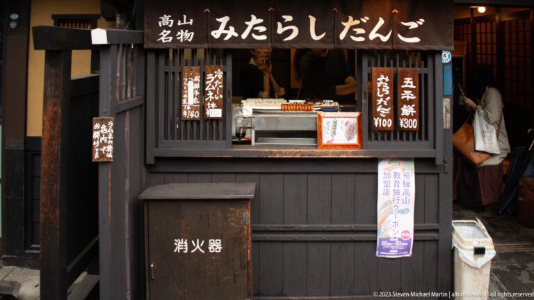 Black wooden street-food stall with signs for mitarashi dango and gohei mochi around a small service counter.