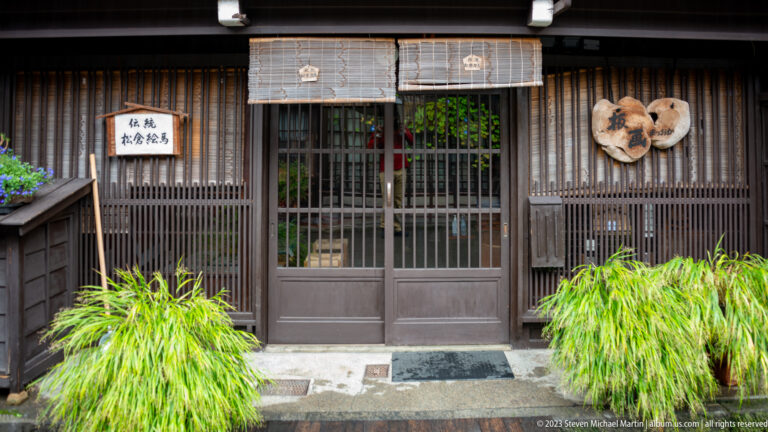 Traditional dark wooden shopfront with lattice doors, bamboo blinds, potted grasses, and a carved wooden emblem.