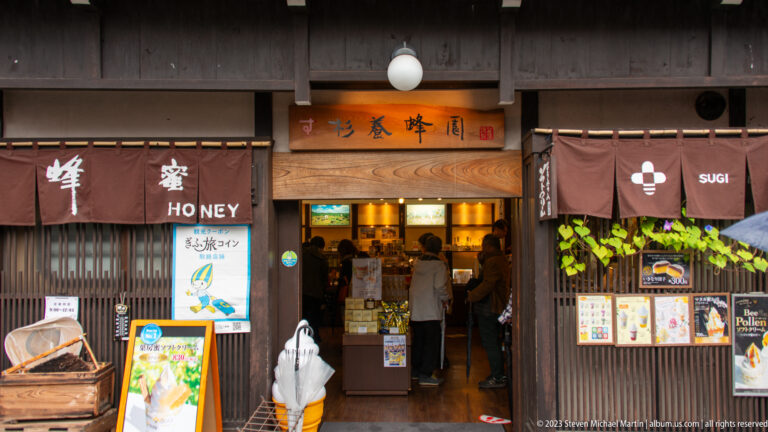 Traditional wooden honey shop with brown noren, honeycomb display, product posters, and a soft-serve model at the entrance.